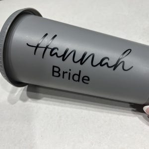 May include: Gray reusable tumbler with a black lid and a black straw. The tumbler has the text "Hannah Bride" written on it.