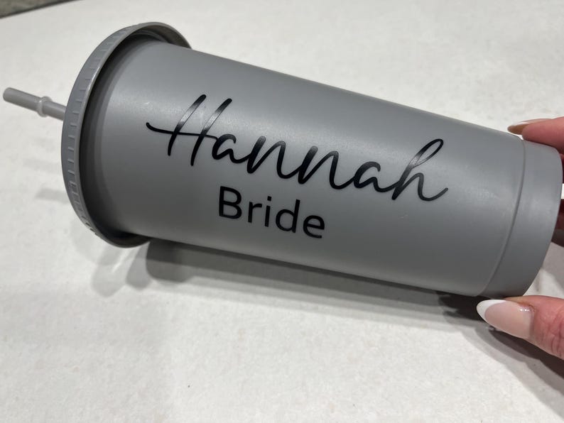 May include: Gray reusable tumbler with a black lid and a black straw. The tumbler has the text "Hannah Bride" written on it.