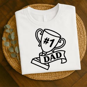 Puede incluir: Camiseta blanca con un gráfico negro de un trofeo, el número 1 y la palabra "DAD" en una pancarta. La camiseta está colocada sobre un salvamanteles redondo tejido.
