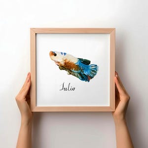 May include: A square wooden frame containing a white print of a betta fish with blue, orange, and white scales. The fish is painted in a textured style, and the name Julio is written in black cursive script underneath the fish illustration.