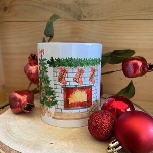 Peut inclure: Une tasse en céramique blanche avec une scène de Noël représentant une cheminée, des chaussettes et un sapin de Noël décoré. La tasse est posée sur une surface en bois avec des boules rouges.