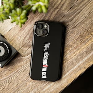 May include: Black phone case featuring the text "Be outstanding out" in white and red. The case is placed on a wooden surface, with a camera and some greenery visible in the background.