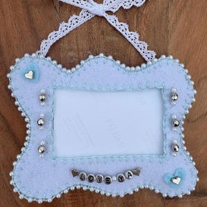 May include: A light blue felt picture frame with a scalloped edge, adorned with faux pearls and silver beads. The frame has a white lace bow at the top and the word "LOVE YA" spelled out in silver beads at the bottom. Two small heart-shaped embellishments are also present.