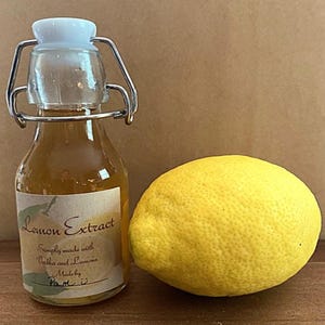 May include: A small glass bottle with a metal clasp closure filled with a light yellow liquid. The bottle has a label that reads "Lemon Extract" and "Simply made with Vodka and Lemons". The label also says "Made by Pam." A whole yellow lemon is next to the bottle.