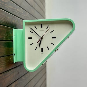 May include: A mint green and white Seiko wall clock with a triangular shape. The clock has black hour markers and hands, with a red second hand. The clock is mounted on a wooden wall.