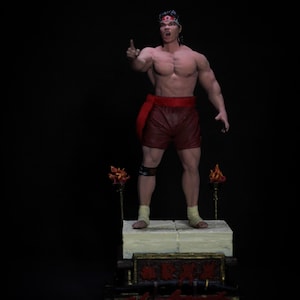 May include: A detailed figurine of a muscular man in red shorts and a headband, pointing upwards. The figure stands on a platform with decorative elements and two torch-like structures. The base has text in red.