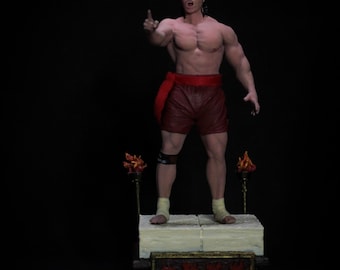 May include: A detailed figurine of a muscular man in red shorts and a headband, pointing upwards. The figure stands on a platform with decorative elements and two torch-like structures. The base has text in red.