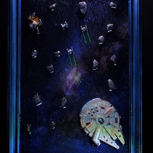 Puede incluir: Una pieza de arte mural 3D enmarcada que muestra la nave espacial Millennium Falcon volando a través de una escena espacial oscura con asteroides y cazas TIE disparando láseres verdes. El fondo muestra una nebulosa y el logo de Star Wars.