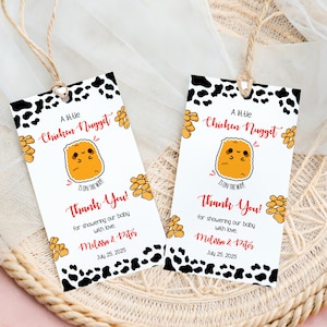 Puede incluir: Dos etiquetas de regalo blancas con un borde de estampado de vaca en blanco y negro y un gráfico de dibujos animados de pepita de pollo. Las etiquetas dicen "A Little Chicken Nugget Is On The Way! Thank You!" e incluyen nombres y una fecha.
