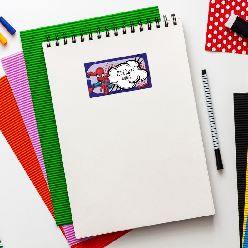 May include: A white notebook with a spiral binding. A sticker with a cartoon superhero character is on the front of the notebook. The sticker says "Peter Jones, Grade 3,  Woof!"
