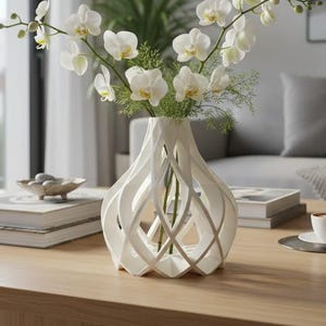Può includere: Un vaso bianco stampato in 3D con una complessa struttura geometrica a reticolo aperto. Il vaso contiene un bouquet di steli di orchidea bianca con fogliame verde. L'oggetto è posizionato su una superficie in legno al centro.