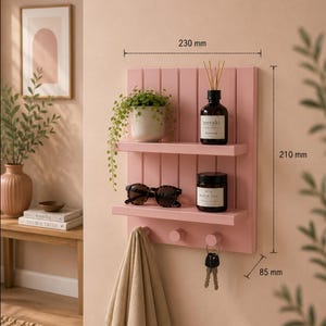 Puede incluir: Un estante de pared de madera rosa con dos repisas horizontales y tres ganchos redondos. La unidad mide 230 mm de ancho, 210 mm de alto y 85 mm de profundidad. Contiene una planta en maceta, un difusor Meraki, gafas de sol, una vela y llaves.