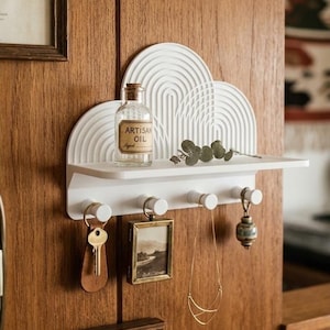 Peut inclure: Une étagère murale blanche dotée d'un panneau arrière semi-circulaire avec un motif d'arche nervuré. L'étagère comprend quatre crochets ronds supportant une clé avec un porte-clés en cuir, un petit cadre photo, un collier doré et un ornement en verre.