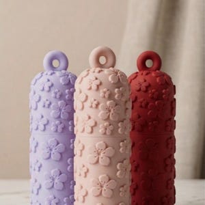 Könnte beinhalten: Drei zylindrische Dekorationsbehälter in Lavendel, Altrosa und Burgunderrot. Jeder hat ein Blumenmuster und einen ringförmigen Griff. Die Behälter bestehen aus einem weichen, matten Material. Höhe ca. 15 cm.