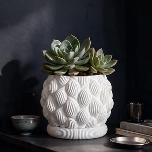 Op de afbeelding: Een witte keramische plantenpot met een gestructureerd, herhalend bladvormig patroon aan de buitenkant. De pot heeft een afgeronde basis en bevat twee groene vetplanten met dikke, vlezige bladeren in een rozetvorm.