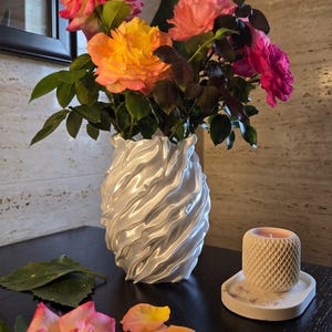 Pode incluir: Um vaso branco com textura ondulada e estriada contém um buquê de rosas cor-de-rosa, laranja e amarelas. Ao lado do vaso, há uma pequena vela cilíndrica com padrão de diamante em uma bandeja oval branca com pétalas espalhadas.