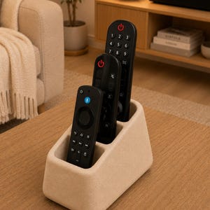 May include: A beige, textured, wedge-shaped remote control holder with three slots, each containing a black remote control. The front remote features a blue microphone icon, while the two taller remotes behind it display red power buttons.