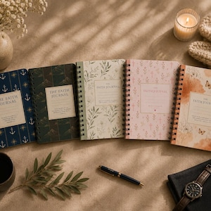 Op de afbeelding: Vijf spiraalgebonden dagboeken getiteld The Faith Journal, elk met unieke omslagontwerpen, waaronder marineblauw met ankers, donkergroen met bergen, crème met bladeren, lichtroze met kruisjes en oranje met bloemen en vlindermotieven.