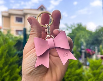 May include: A pink leather bow keychain with a gold ring and clasp. The keychain has the letters "eb" embossed on the bow.