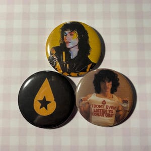 May include: Three circular pin-back buttons. One features a person with dark curly hair against a yellow background. Another is black with a yellow teardrop shape containing a black star. The third shows a person wearing a white tank top with the text I DONT EVEN LISTEN TO CONAN GRAY.