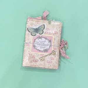 May include: A pink and cream patterned journal with a butterfly sticker, a key sticker, and the text "The world is full of wonderful things!"