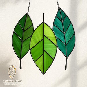 Può includere: Tre ornamenti a forma di foglia in vetro colorato nelle tonalità del verde, ciascuno con una cornice e uno stelo in metallo nero. Le foglie sono sospese da una catena. Il logo "CHRYALUME STAINED GLASS PATTERNS" è in basso.