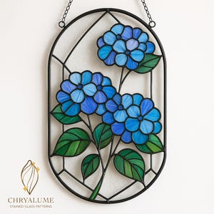 Peut inclure: Panneau en vitrail représentant des fleurs d'hortensia bleues avec des feuilles vertes. Les fleurs sont dans des tons de bleu, et les feuilles sont vertes. Le panneau est encadré de métal noir et comprend le texte "CHRYALUME STAINED GLASS PATTERNS".
