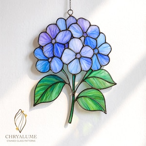 Könnte beinhalten: Buntglas-Hortensienblume mit blauen, lila und weißen Blütenblättern, grünen Blättern und einem grünen Stiel. Das Kunstwerk ist mit dunklem Metall gerahmt und hängt an einer Kette. Das Logo "CHRYALUME STAINED GLASS PATTERNS" befindet sich unten.