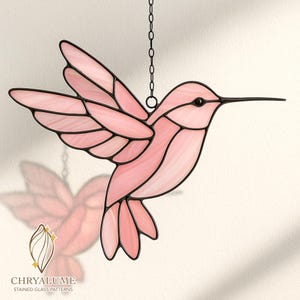 Könnte beinhalten: Ein rosafarbener Glas-Hummingbird mit schwarzen Umrissen und einem langen Schnabel hängt an einer Kette. Die Flügel des Vogels sind im Flug ausgebreitet, und das Glas hat eine durchscheinende Qualität. Das Logo "CHRYALUME STAINED GLASS PATTERNS" befindet sich unten.