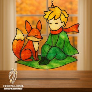 May include: Stained glass art piece depicting the Little Prince and a fox. The artwork features a young boy in a green outfit and red scarf, seated next to an orange fox on a green surface. The piece is suspended by a chain.