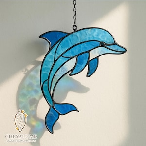 Peut inclure: Un dauphin en vitrail aux tons bleus, avec un contour noir et une chaîne pour l'accrocher. Le dauphin est représenté en train de sauter, avec un aspect texturé. Le logo "CHRYALUME STAINED GLASS PATTERNS" est visible en bas.
