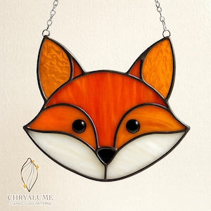 Puede incluir: Cabeza de zorro de vidriera con detalles en naranja, blanco y negro. El zorro tiene nariz y ojos negros, con orejas naranjas. Cuelga de una cadena. El logotipo "CHRYALUME STAINED GLASS PATTERNS" está en la parte inferior.