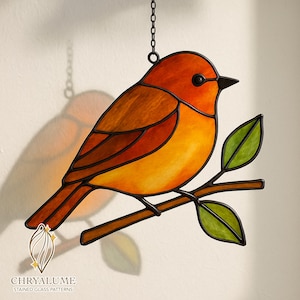 May include: A stained glass bird suncatcher in orange, brown, and yellow, perched on a branch with green leaves. The bird is suspended by a chain. The logo "CHRYALUME STAINED GLASS PATTERNS" is at the bottom.