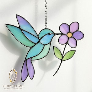 May include: Stained glass hummingbird with a flower. The bird features shades of blue, green, and purple, with a black outline. The flower is purple with a yellow center. The piece hangs from a black chain. The logo "CHRYALUME STAINED GLASS PATTERNS" is at the bottom.