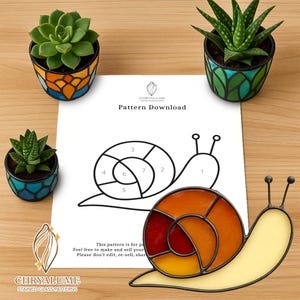 May include: A stained glass snail with a yellow body and an orange and red shell sits next to a pattern download. The image also features three small potted succulents with colorful, geometric pots. The text on the paper reads "Pattern Download".