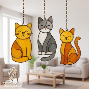Könnte beinhalten: Drei Glasmalerei-Katzenornamente in Gelb, Grau und Orange, jeweils an einer Kette aufgehängt. Die Katzen haben lächelnde Gesichter und sitzen. Der Hintergrund zeigt ein Wohnzimmer mit Sofa und Couchtisch.