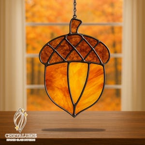 Può includere: Acchiappasole a forma di ghianda in vetro colorato con cornice in metallo scuro. La parte superiore della ghianda è marrone e il corpo è un mix di arancione e giallo. L'acchiappasole è appeso a una catena.