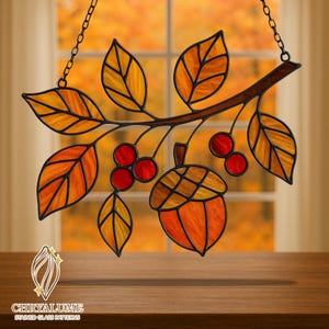 May include: Stained glass autumn-themed art piece featuring a branch with leaves, berries, and an acorn. The leaves are in shades of orange and yellow, with red berries and an orange acorn. The piece is framed with black metal and hangs from a chain. The text "CHRYALUME STAINED GLASS PATTERNS" is at the bottom.