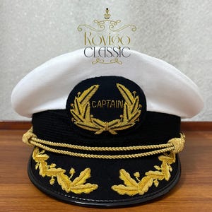 May include: A white captain's hat with a black band and gold embroidery. The hat features the word "CAPTAIN" in gold lettering, surrounded by a gold wreath. The hat has gold rope detailing and gold leaf embroidery on the brim.