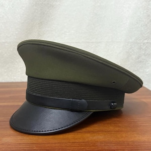 May include: A green military-style cap with a black leather brim. The cap has a black band with a decorative stitched detail.