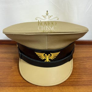 Puede incluir: Una gorra de estilo militar en color beige con una banda negra y un emblema de águila bicéfala bordado en oro. La gorra tiene una visera beige e incluye el texto Rovigo CLASSIC impreso en dorado sobre la parte superior de la corona.