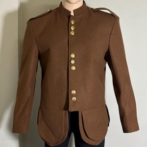 Pode incluir: Uma jaqueta de lã marrom com gola mandarim e botões dourados. A jaqueta tem um estilo longo e ajustado com cintura alta e saia levemente evasê.