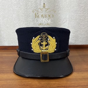 Op de afbeelding: Marineblauwe militaire hoed met een zwart vizier en een gouden embleem. Het embleem toont een gouden anker omringd door gouden bladeren. De hoed heeft een zwarte leren riem en een gouden gesp. De tekst "Rovigo Classic" is zichtbaar.