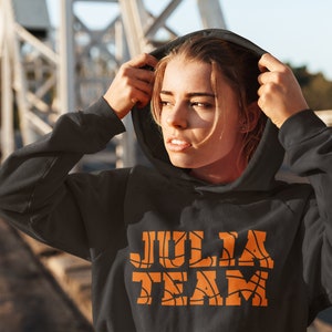 以下が含まれることがあります： バスケットボール柄のオレンジ色の文字で「JULIA TEAM」と書かれた黒いフード付きスウェットシャツ。テキストはスウェットシャツの中央に配置されています。