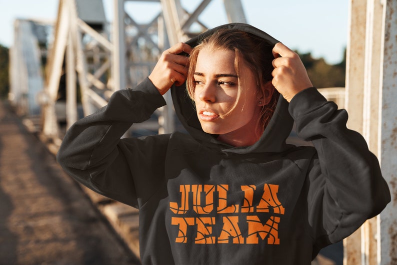 以下が含まれることがあります： バスケットボール柄のオレンジ色の文字で「JULIA TEAM」と書かれた黒いフード付きスウェットシャツ。テキストはスウェットシャツの中央に配置されています。