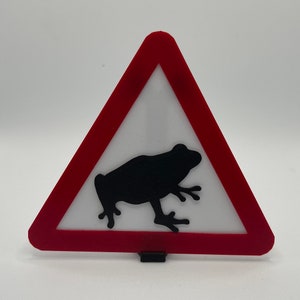 Puede incluir: Una señal de advertencia triangular roja con una silueta negra de una rana en su interior. La señal es una advertencia para estar atento a las ranas en la zona.