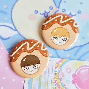 Pode incluir: Dois botões redondos, cor de bronzeado claro, com uma ilustração de desenho animado de um personagem dentro de um design estilo takoyaki. Os botões têm detalhes marrons e brancos, e o personagem tem um rosto sorridente.