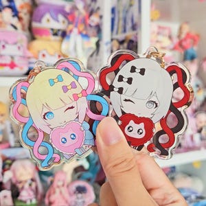 May include: Two colorful anime character keychains with a heart-shaped design. The keychain on the left features a girl with blonde hair and blue eyes, wearing a pink and blue outfit. The keychain on the right features a girl with white hair and blue eyes, wearing a red and black outfit.