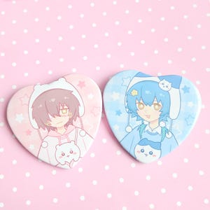 Może przedstawiać: Dwie przypinki w kształcie serca z postaciami w stylu anime. Jedna przypinka ma różowe tło i postać z brązowymi włosami, druga niebieskie tło i postać z niebieskimi włosami. Obie przypinki mają uroczy zwierzęcy design.