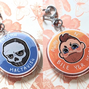 May include: Two circular acrylic keychains with silver-tone metal clasps. The left keychain features a skull mask illustration on a blue background with the text English Mactavish. The right keychain shows a cartoon character on an orange background with the text Away N' Bile Yer Heid!